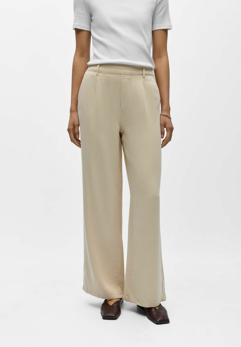 Femme portant un pantalon beige à jambes larges, un haut blanc à manches courtes, et des chaussures à bout carré de couleur brun foncé, debout devant un fond uni.