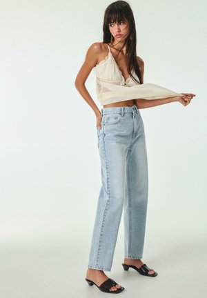 Frau trägt ein cremefarbenes, mit Spitze verziertes Camisole, hellblaue hoch taillierte Jeans und schwarze Sandalen mit Absatz, steht vor einem schlichten weißen Hintergrund.