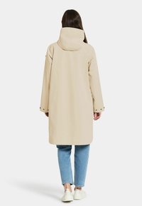 Beige waterdichte lange jas met een capuchon, voorzien van drukknoopsluitingen op de mouwen en een ontspannen pasvorm. Gecombineerd met lichtblauwe jeans en witte schoenen.