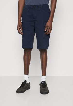 Shorts cargo navy en mélange de coton, dotés de poches latérales et d'une coupe décontractée. Portés avec des baskets noires et vertes, et des chaussettes blanches courtes.