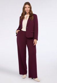 Woman standing wearing a plum-colored pantsuit with a white blouse and cream ankle boots on a plain light background.