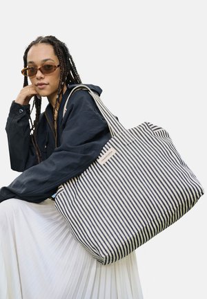 Personne aux cheveux tressés et lunettes de soleil reposant le menton sur la main, portant une veste foncée et une jupe blanche, portant un grand sac cabas rayé bleu marine et blanc.