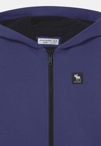 Sudadera con capucha morada con cremallera, forrada con polar negro, que presenta un logo cuadrado negro con un diseño de alce. Etiqueta de talla visible en el interior.