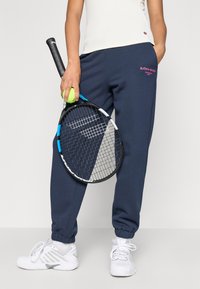 Navy zils sporta bikses ar elastīgām piedurknēm, valkājot baltas sporta kurpes. Tur rokās tenisa raketi un dzeltenu tenisa bumbu. Vienkāršs dizains.