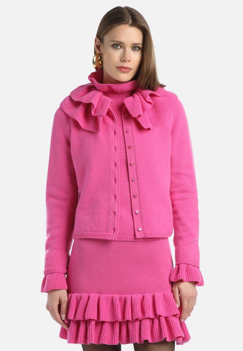 Cárdigan de punto rosa con cuello volante, cierre de botones y falda a juego con volante. Textura suave con puños y dobladillo acanalados.