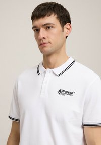 Biała koszulka polo z teksturowanej tkaniny, z czarnym haftowanym logo oraz przyciskiem na dwa guziki z czarnymi akcentami na kołnierzyku.