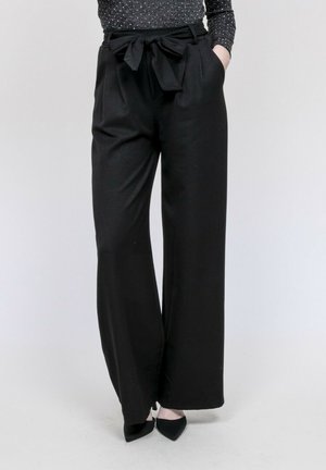 Femme portant un pantalon large noir taille haute avec une ceinture nouée, associé à un haut à manches longues à pois et des chaussures noires à bouts pointus.