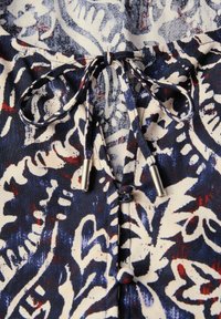 Detail látky v navy a krémovém vzoru se zavazováním na mašli, které má stříbrné konce. Design zahrnuje točité květinové motivy.
