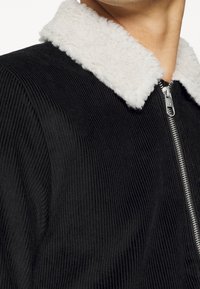 Svart kordflöjeljacka med en vit shearlingkrage, med en framficka med dragkedja och ribbad textur i tyget.