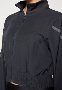 Černá bunda na zip vyrobená z hladké, lehké látky. Má límec, elastický spodní okraj a reflexní pruhy na ramenou.