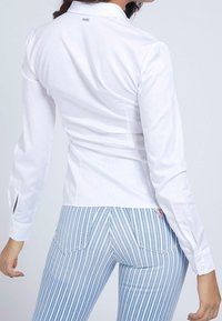 Chemise blanche à manches longues avec une coupe ajustée, dotée de poignets boutonnés et d'une petite étiquette de logo au dos. Associée à un pantalon rayé bleu et blanc.