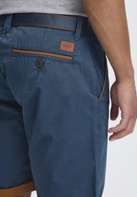 Marineblå shorts i et lett stoff med oransje-brun aksent på lommene og midjebåndet. Har et merke med "BLEND" logo.