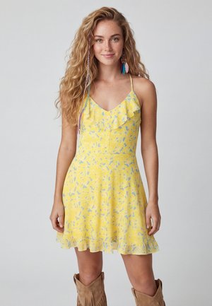 Jonge vrouw met lang krullend haar draagt een gele zomerse jurk met bloemenprint en ruches, gecombineerd met bruine laarzen met franjes, staande tegen een effen achtergrond.
