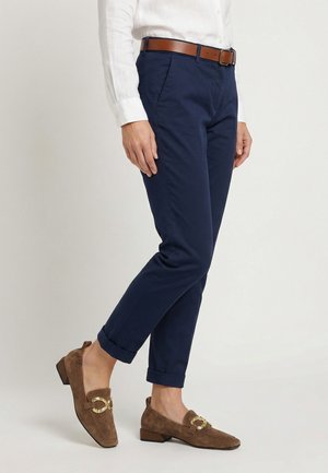 Person trägt marineblaue, umgeschlagene Hose, ein in die Hose gestecktes weißes Hemd, einen braunen Gürtel und braune Wildleder-Loafers mit goldenen Schnallenakzenten.