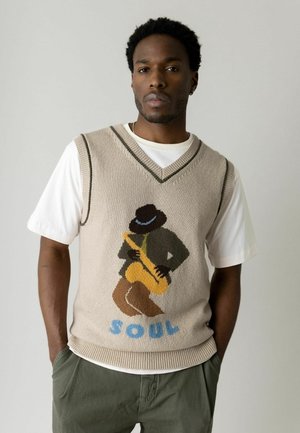 Hombre con chaleco de punto beige sobre camiseta blanca, el chaleco muestra a un músico de jazz tocando el saxofón con la palabra "SOUL" debajo.