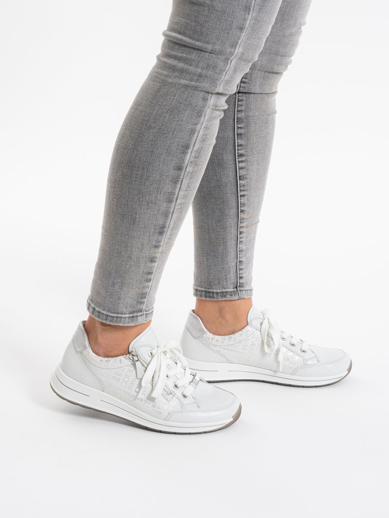 Gambe con jeans aderenti grigio chiaro e scarpe da ginnastica bianche con lacci e cerniere laterali, in piedi su uno sfondo bianco.