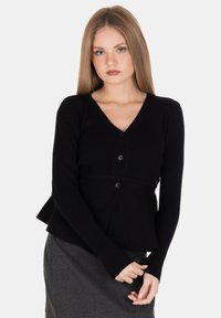 Giovane donna con lunghi capelli biondi, che indossa un cardigan nero con bottoni e una gonna grigia, in piedi con le mani dolcemente unite davanti.