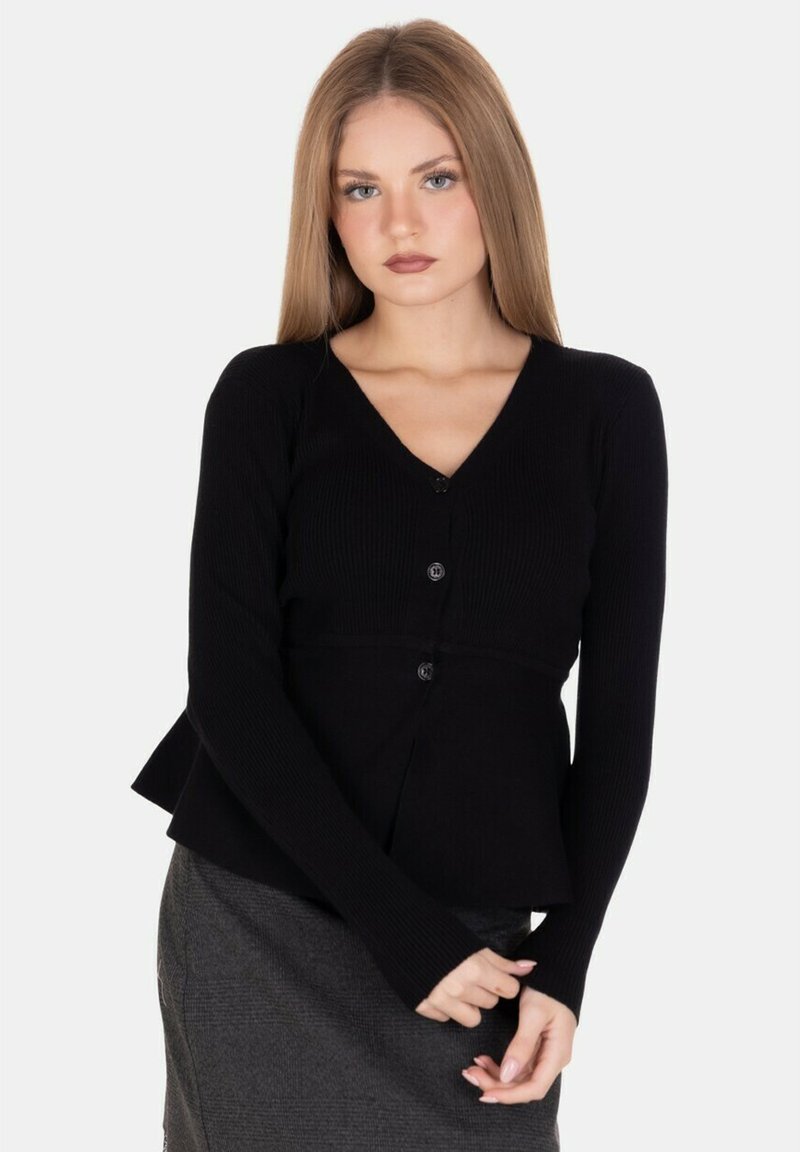 Giovane donna con lunghi capelli biondi, che indossa un cardigan nero con bottoni e una gonna grigia, in piedi con le mani dolcemente unite davanti.