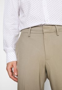 Beige maßgeschneiderte Hose mit glatter Textur, die eine figurbetonte Silhouette und eine sichtbare Mittelfalte auf der Vorderseite aufweist. Kombiniert mit einem leichten weißen Hemd, das mit einem kleinen schwarzen geometrischen Muster verziert ist.