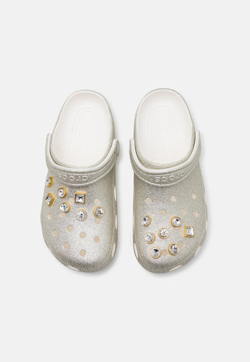 Metallic silberne Clogs mit runden Belüftungsöffnungen und dekorativen Edelsteinen in verschiedenen Formen, einer strukturierten Oberseite und einem stabilen Riemen.