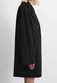 Abrigo de lana negra con un corte relajado, collar muescado, dos bolsillos delanteros y cierres de botón. Textura suave con un diseño sin costuras.