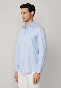 Camisa de manga larga azul claro con rayas blancas verticales, cuello abotonado y dobladillo redondeado. La tela parece suave y ligera.