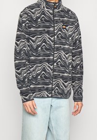 Fleece'owa kurtka z falistym abstrakcyjnym wzorem w kolorze granatowym, szarym i białym, z zapięciem na zamek z przodu i małym logo na piersi.