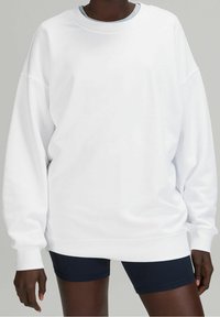 Felpa bianca oversize con scollo rotondo e maniche lunghe, caratterizzata da una texture morbida e polsini a coste. Indossata sopra dei pantaloni corti blu navy.