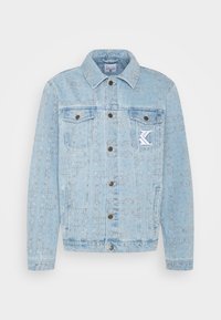 Light blue denim jacket with intricate embossed patterns, two chest pockets, and metal button closures. Features a white logo patch.