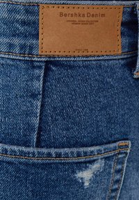 Jeans in denim blu con etichetta in pelle incisa "Bershka Denim." Presentano una texture usurata e dettagli sulle tasche posteriori.