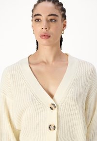 Crème-kleurige gebreide cardigan met een V-hals, voorzien van drie grote ronde knopen en een losse pasvorm. Textuurstof met een ribbelpatroon.