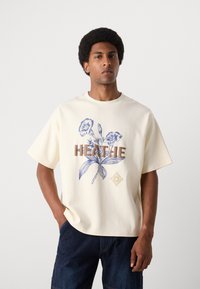 Cremefarvet t-shirt med korte ærmer, der har blå blomster grafik og brune bogstaver. Glat tekstur med en afslappet pasform.