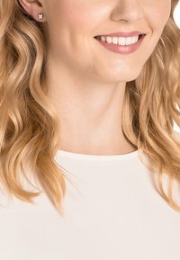 Brincos de prato quadrados com um acabamento polido, usados por uma pessoa com cabelo loiro ondulado, combinados com uma blusa de cor clara.