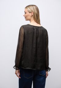 Blusa nera trasparente con motivo a stampa leopardata, maniche lunghe e polsini elastici. Scollo a V sul retro, texture morbida, tessuto leggero.