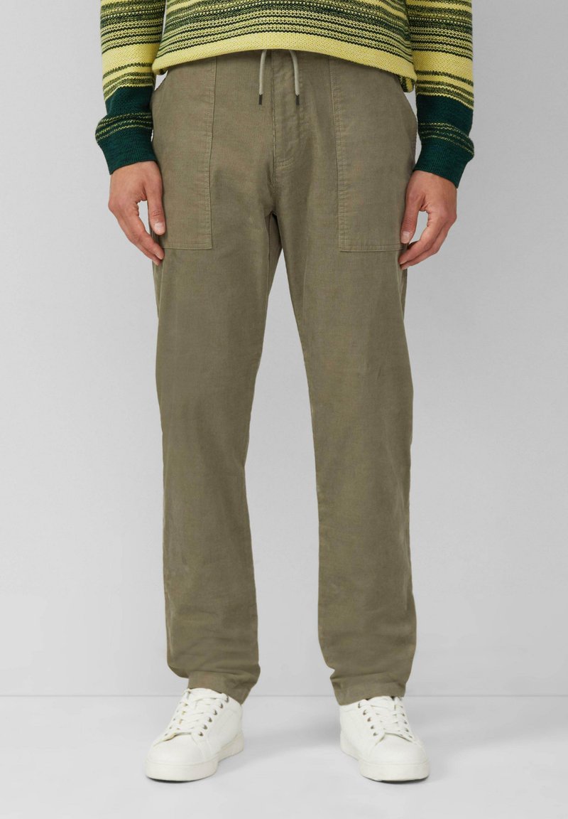 Kaki kleurige broek van textuurstof, met een elastische tailleband en twee grote zijzakken. Draagt een paar witte sneakers.