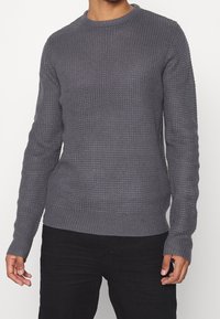 Mężczyzna ubrany w długi rękaw, teksturowany szary sweter dzianinowy i czarne spodnie, stojący z rękami swobodnie wzdłuż ciała.