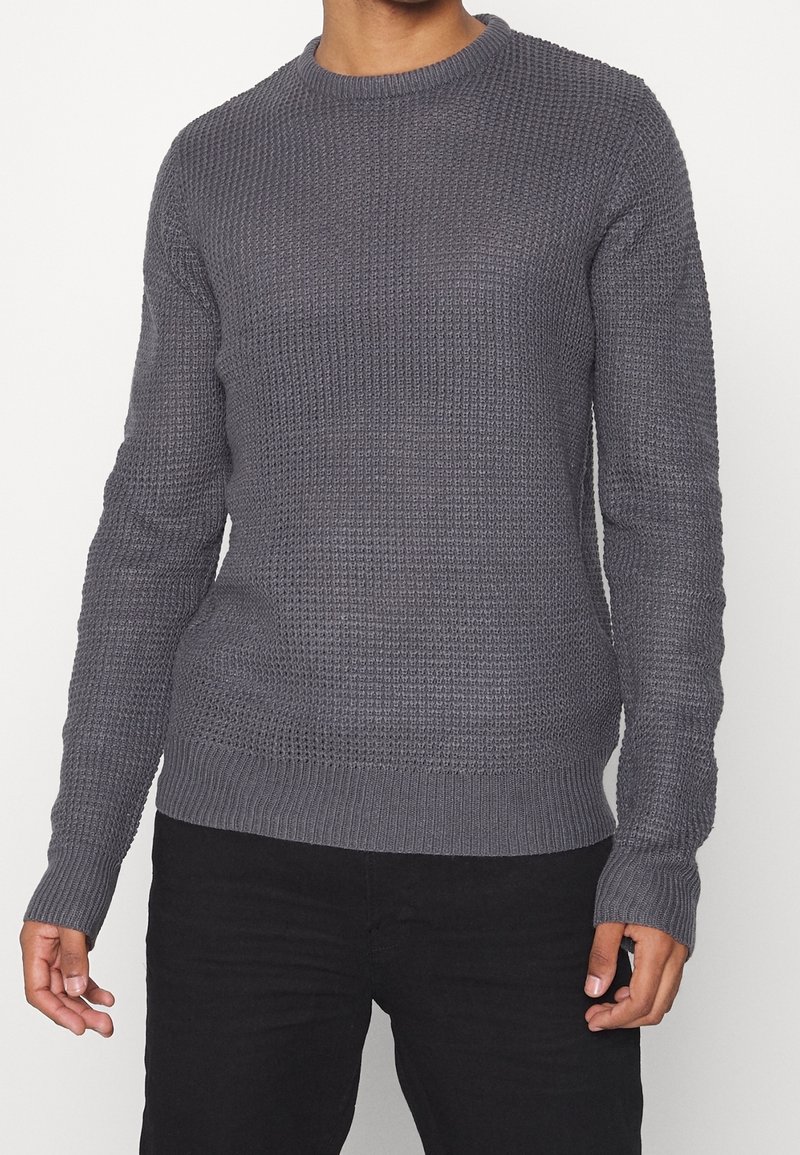 Mężczyzna ubrany w długi rękaw, teksturowany szary sweter dzianinowy i czarne spodnie, stojący z rękami swobodnie wzdłuż ciała.