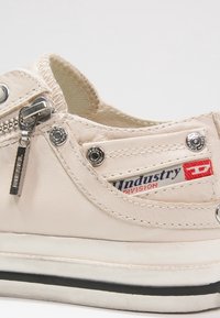 Sneaker in pelle crema dal profilo basso, caratterizzata da una cerniera laterale, accessori in metallo argentato e una suola nera a contrasto.