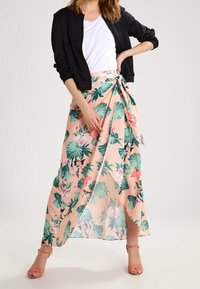 Žena v černé bundě, bílé halence a broskvovém květinovém zavinovacím sukně se zelenými listy a růžovými květy, doplněná o broskvové lodičky.