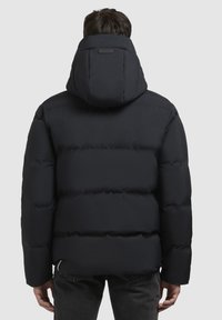 Zwarte gevoerde jas met een capuchon, voorzien van gestikte secties, een gladde textuur en een logo-tag op de achterkant. Draagt een donkere spijkerbroek.