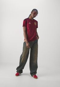 Sarkana futbola krekls ar īsām piedurknēm, dimanta tekstūra un zelta akcenti, apvienots ar brīviem ziliem džinsiem un sarkaniem treneriem. Valkā skaidras saulesbrilles.