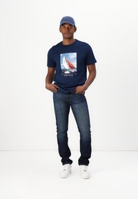 Mørkeblå bomulls-T-skjorte med seilgrafikk, kombinert med mørkeblå jeans og hvite joggesko, komplettert med en lys blå caps.