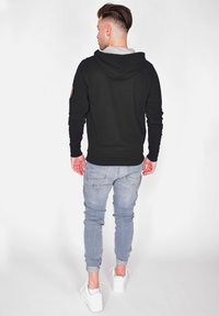 Svart luvtröja med grå innervfodring, ribbade muddar och en logotypapplikation på ärmen, i kombination med ljusblå tapered jeans och vita sneakers.