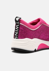 Zapatilla de deporte rosa con brillantes, con parte superior de tela suave, entresuela blanca, suela de goma negra y un tirador negro con un detalle de logo blanco.