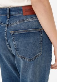 Blå denimjeans med medelhögt midja, försedda med en brun läderetikett märkt "ONLY" och en enda bakficka med kontrasterande sömnad.