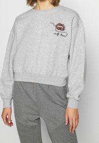 Sudadera gris recortada con mangas anchas, con un diseño de labios rosa y marrón y el texto "amor propio" en negro cerca de la parte superior izquierda.