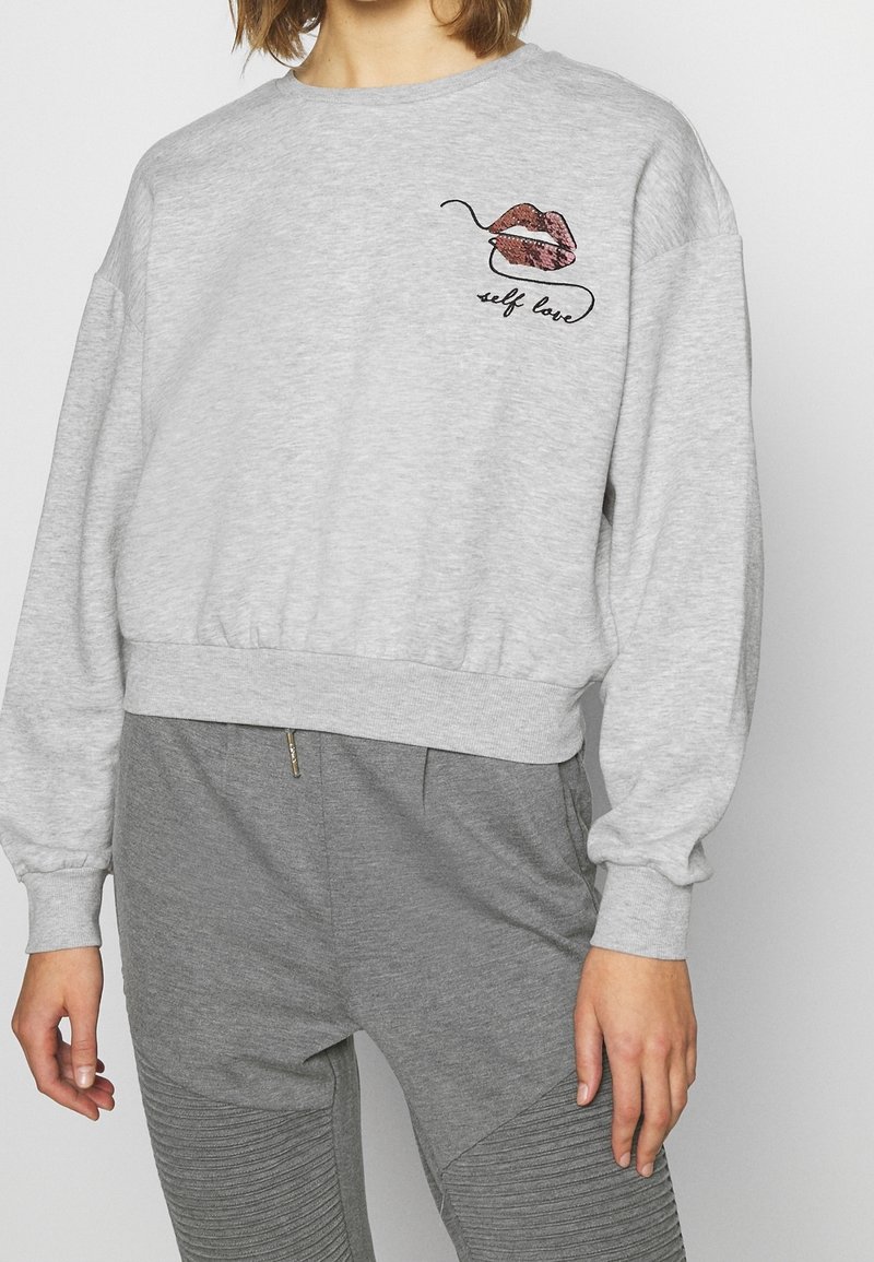 Sudadera gris recortada con mangas anchas, con un diseño de labios rosa y marrón y el texto "amor propio" en negro cerca de la parte superior izquierda.