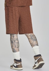 Braune gemusterte Shorts aus weichem Stoff, mit einem dezenten Logo-Design; getragen mit weißen Socken und braunen High-Top-Sneakern.