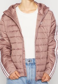 Puchowa kurtka w miękkim mauve z zapięciem na zamek, kapturem i białymi paskami wzdłuż rękawów. Posiada pikowaną strukturę i lekką tkaninę.