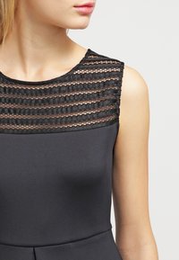 Robe noire avec un corsage ajusté, présentant un col en maille rayée. Texture lisse avec un éclat subtil, mettant en valeur une silhouette élancée.